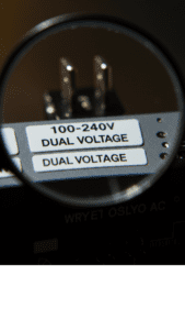 Vista detalhada em macro de etiqueta de um aparelho eletrônico, com foco seletivo destacando as indicações de voltagem "100-240V" e "Bivolt", com uma lupa ampliando detalhes importantes.